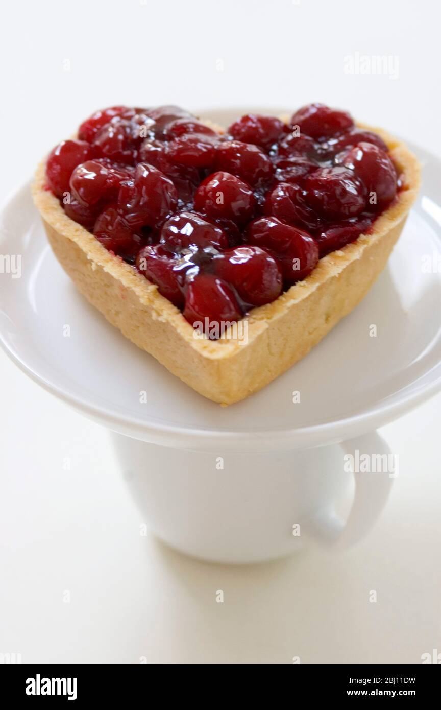 Crostata di ciliegia a forma di cuore su piattino bianco equilibrante su tazza - Foto Stock