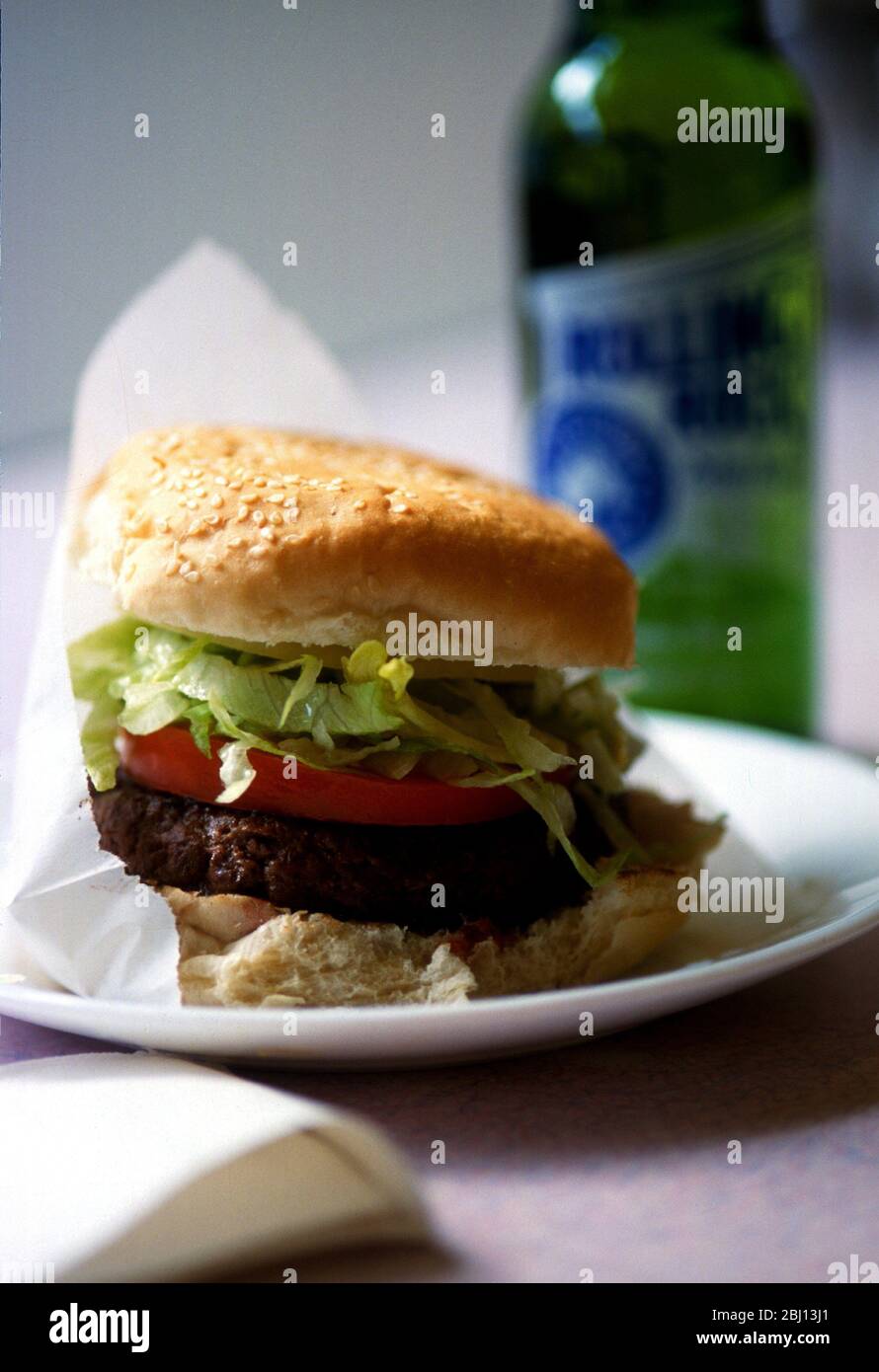 Hamburger americano classico con pomodoro e lattuga grattugiata in panino di sesamo servito in sacchetto di carta su piatto bianco con bottiglia di birra Rolling Rock, a forma Foto Stock