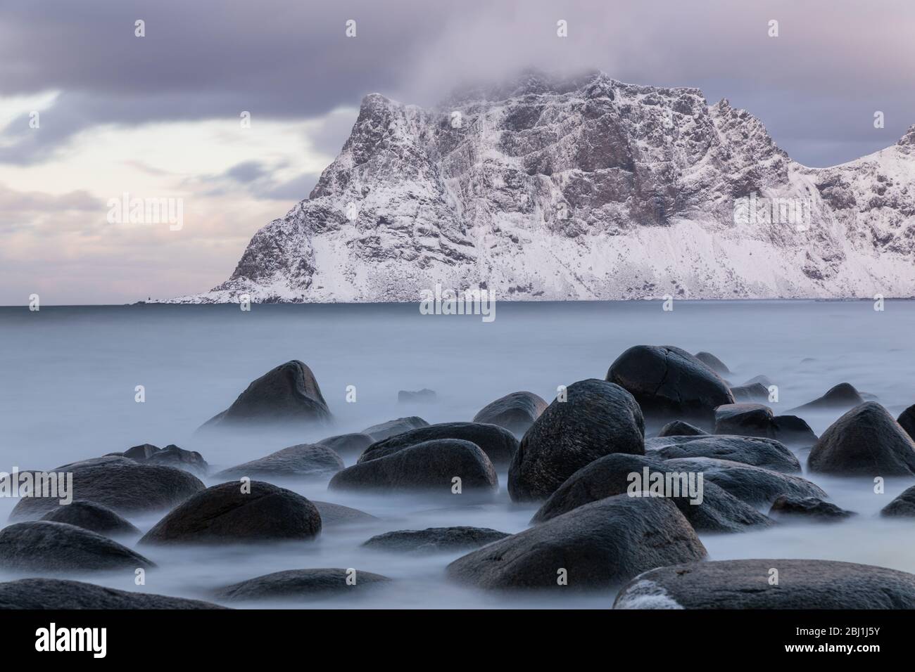 Cattura a lunga esposizione della spiaggia di Uttakleiv, Lofoten, Norvegia. Mare e rocce soffici. Neve coperta montagna sullo sfondo. Calmo scenario invernale. Foto Stock