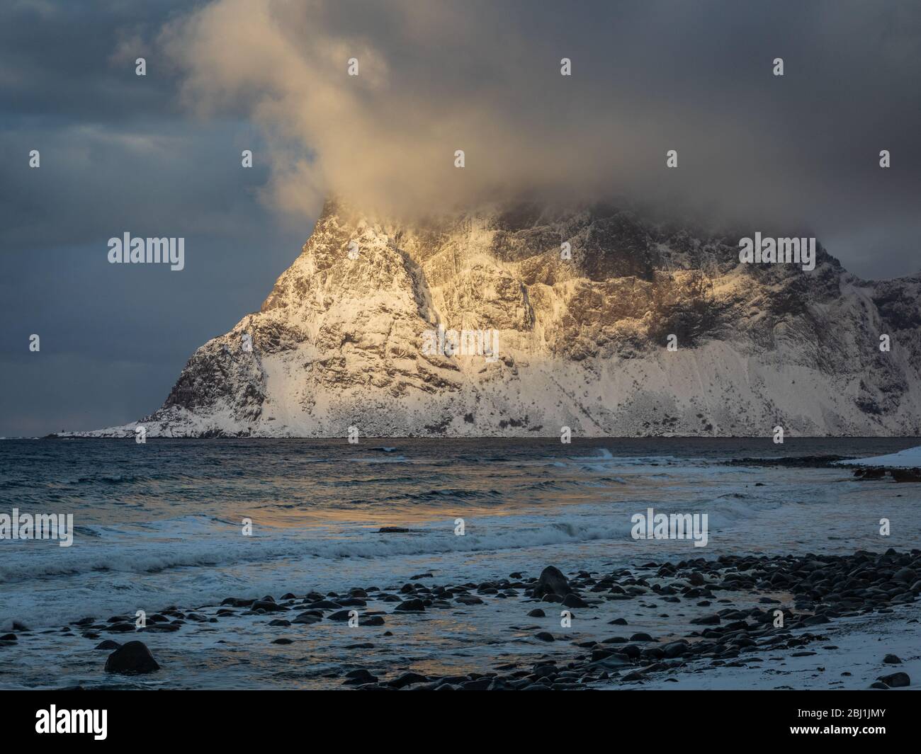 Tramonto sulla spiaggia di Uttakleiv, Lofoten, Norvegia. Ultima luce del sole luce neve coperta montagna e la sua cima sapeared in nuvole tempestose Foto Stock
