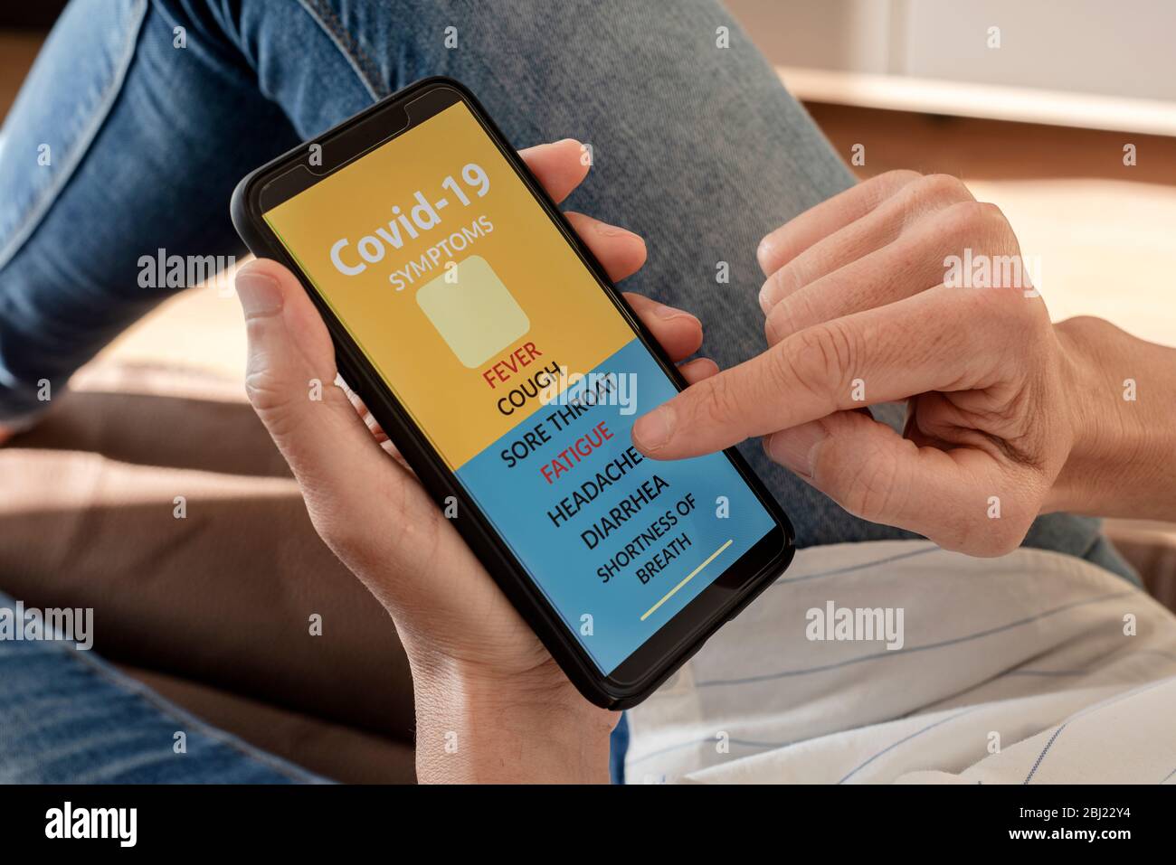 primo piano di un giovane caucasico, comodamente sdraiato su un divano, controllando alcuni dei sintomi del covid-19 in una app simulata nel suo smartphone Foto Stock