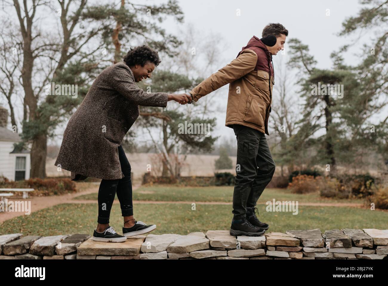 Un uomo bianco che tiene le mani con una donna nera mentre camminano lungo la cima di un muro in un giardino. Foto Stock