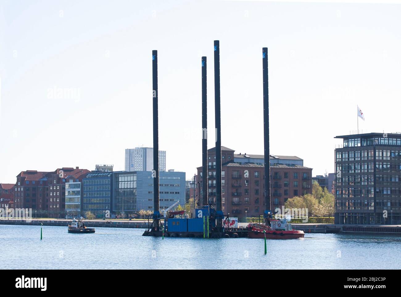 Rimorchiatore trainante di una grande chiatta attraverso un porto urbano. La macchina sulla chiatta è per studi geotecnici. Moderni edifici residenziali nel backgrou Foto Stock