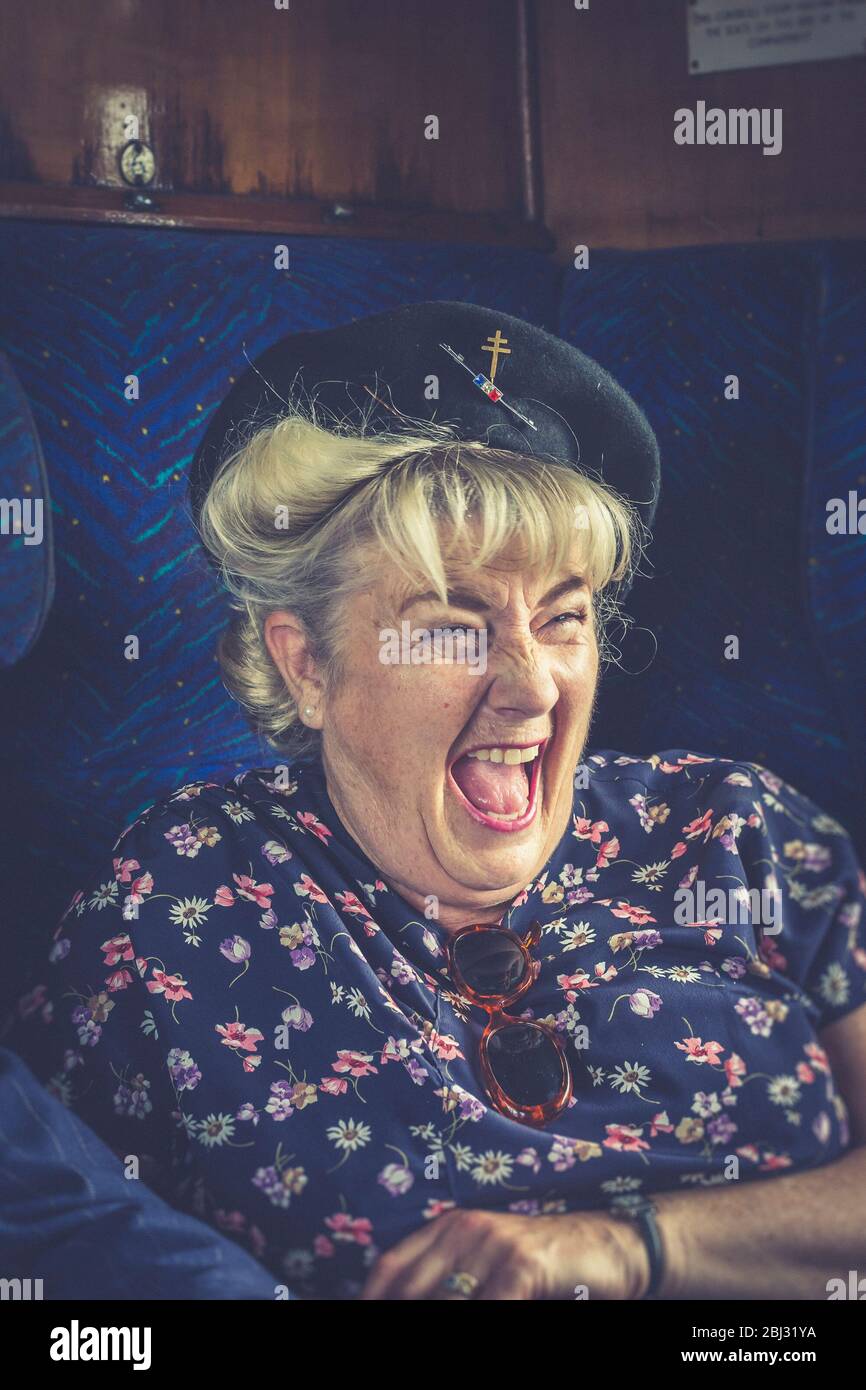Primo piano ritratto di ridere 1940s donna a bordo del treno a vapore del Regno Unito d'epoca, isolato all'interno di una carrozza ferroviaria d'epoca, WW2 evento ferroviario storico. Foto Stock