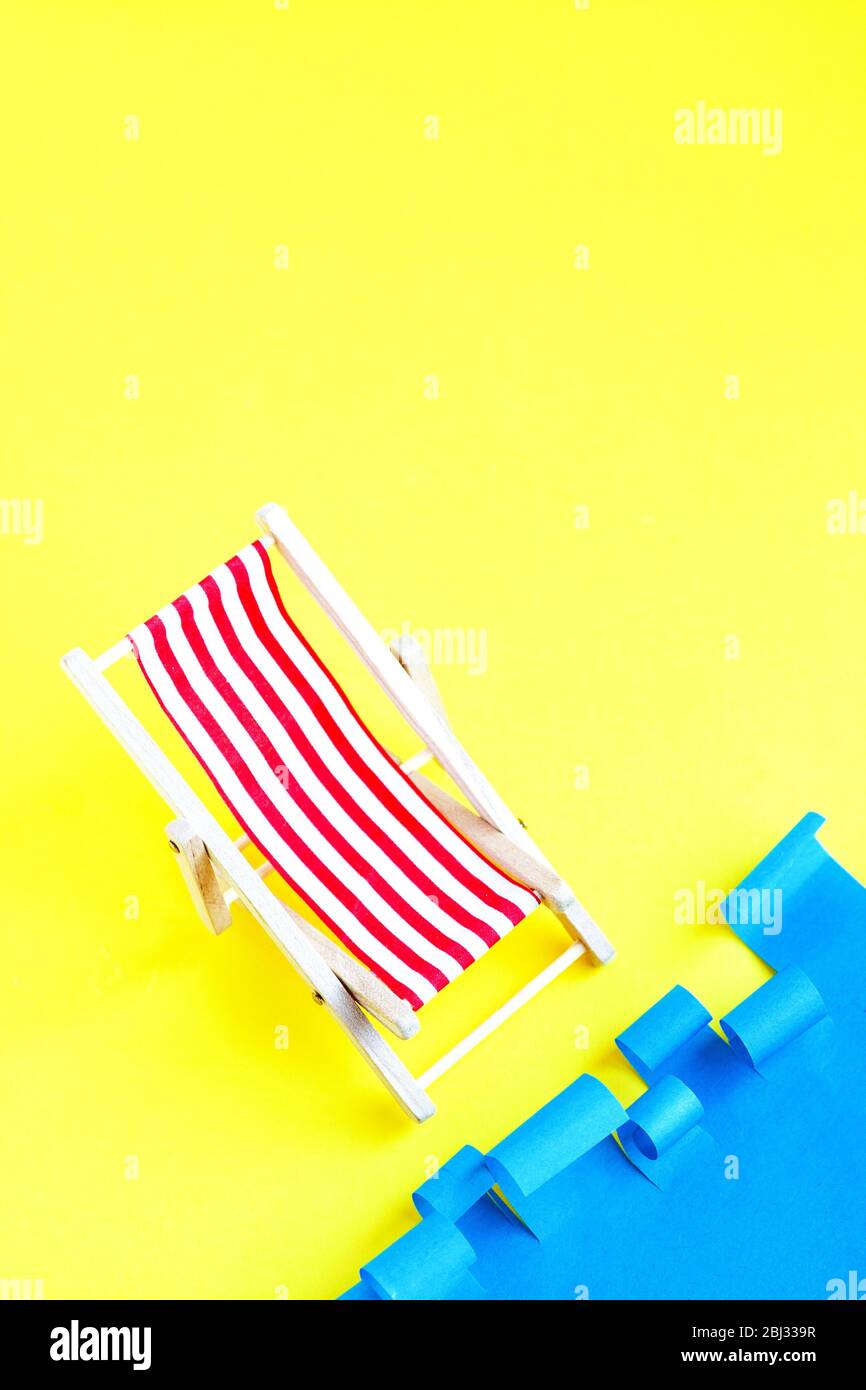 Sedia a sdraio rossa a righe vicino al mare. Sedia reclinabile giocattolo su sfondo blu e giallo. Concetto riposo estivo. Vacanze. Foto Stock