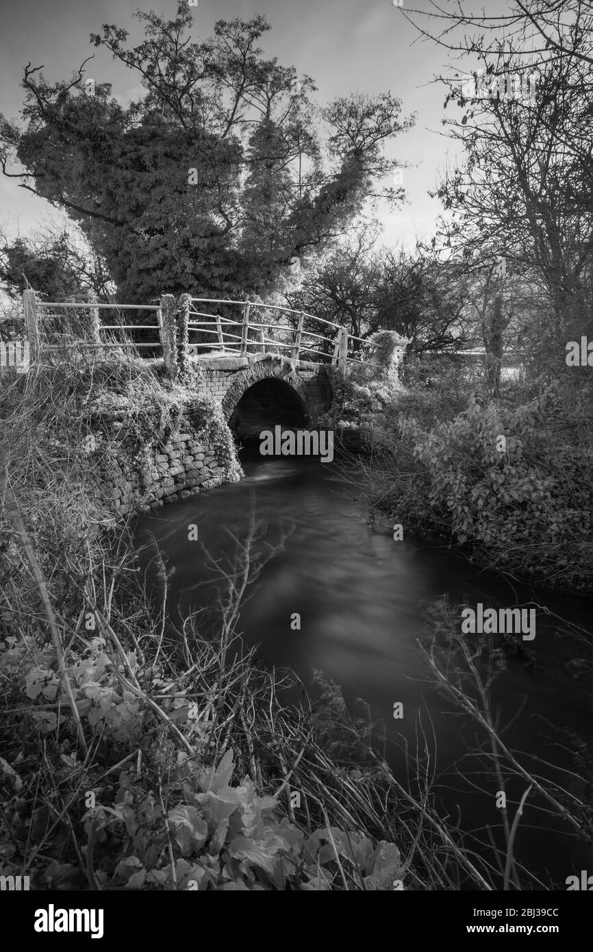Ponte pedonale in mattoni sul fiume Stiffkey, Norfolk, Inghilterra. Immagine a infrarossi in bianco e nero. Foto Stock