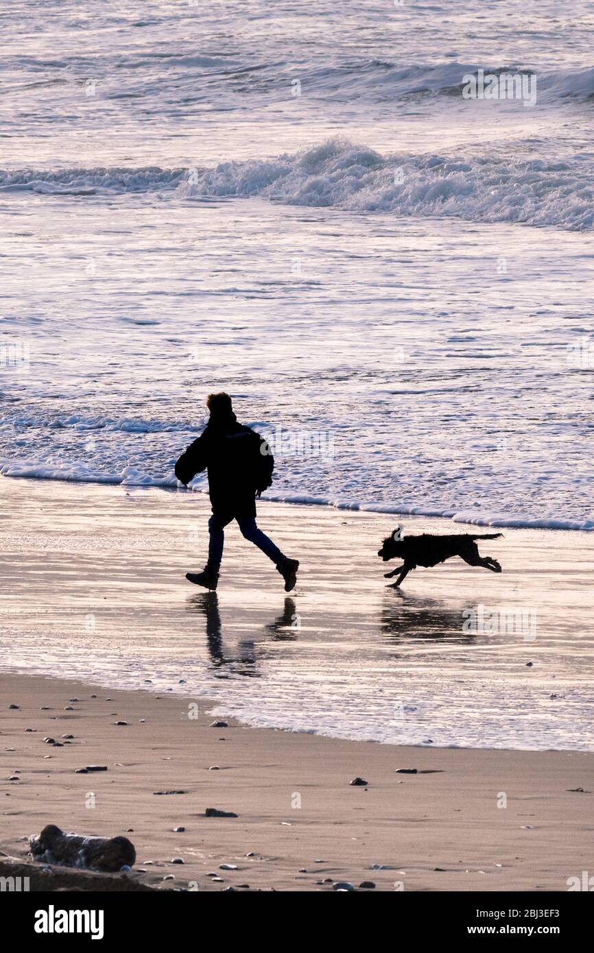 La silhouette di un giovane ragazzo giocando con il suo cane nella luce della sera su Fistral Beach in Newquay in Cornovaglia. Foto Stock