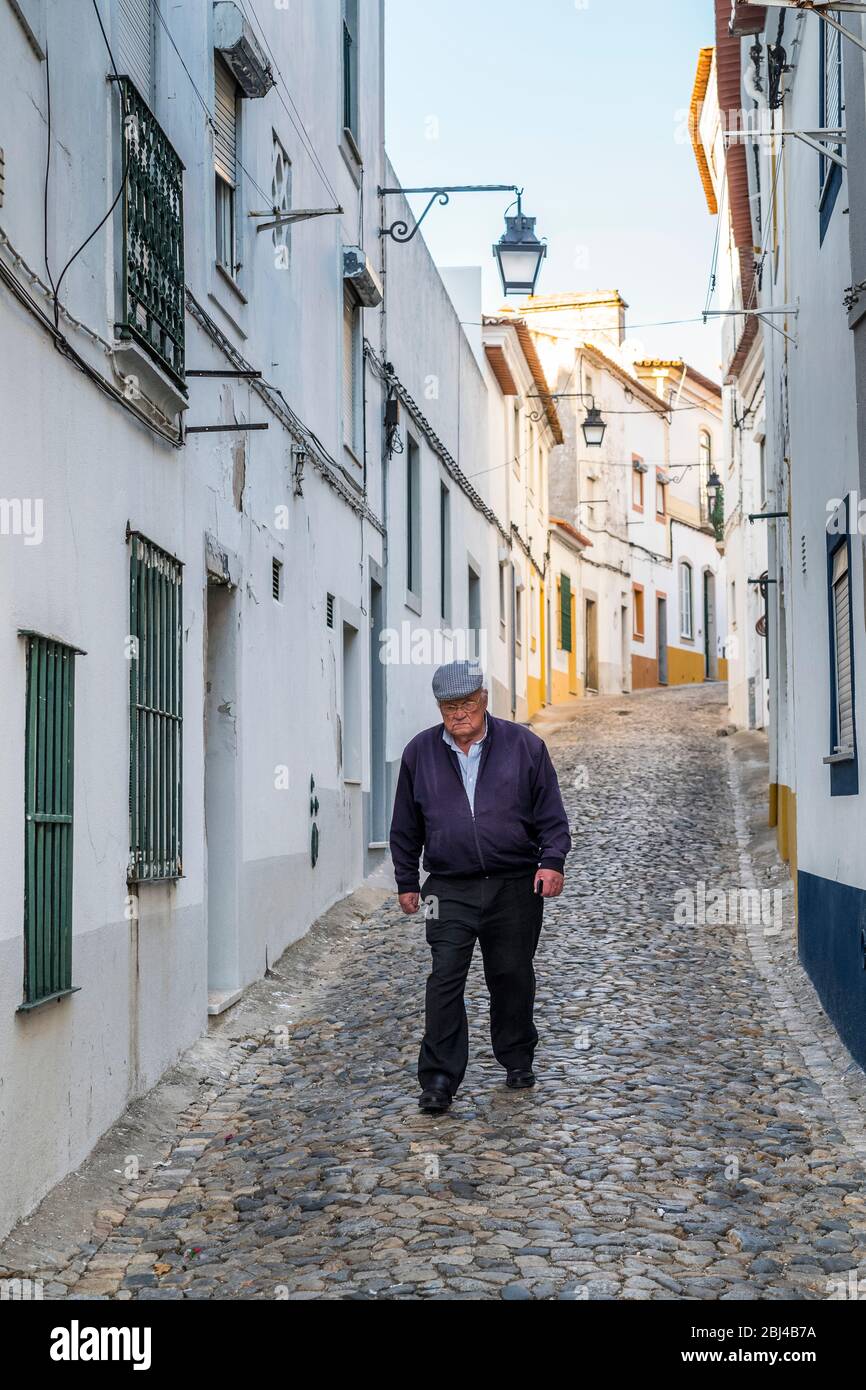 Uomo anziano in piano CAP camminare in tipico scenario di strada di case bianche e gialle, lanterne e strada stretta ciottoli a Evora, Portogallo Foto Stock