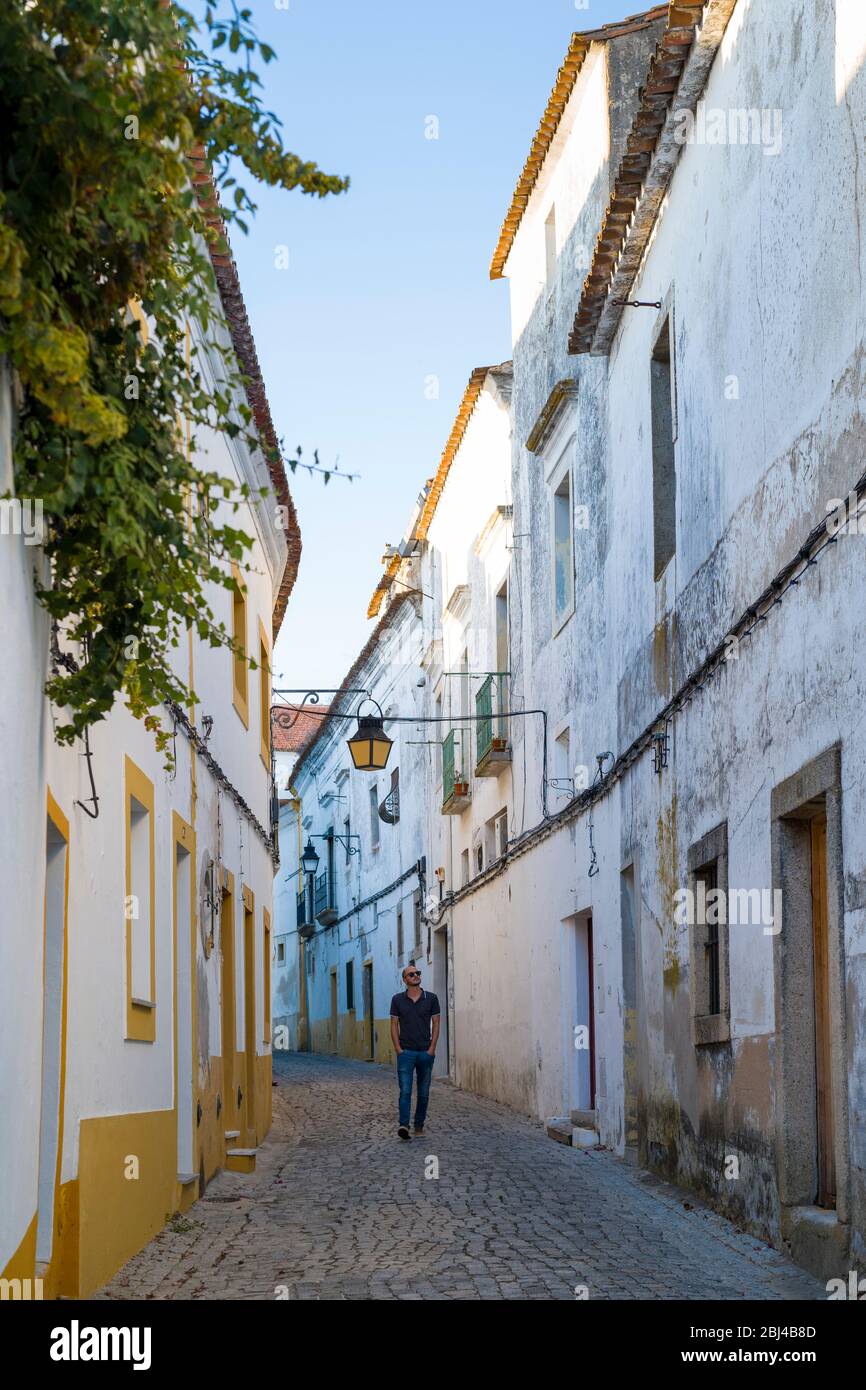 Giovane uomo, mani in tasca, in tipico scenario di strada di case bianche e gialle, lanterne e strada ciottoli stretta, Evora, Portogallo Foto Stock