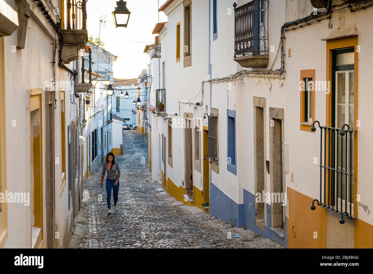 Giovane donna in abiti casual camminando in strada ciottoli passando tipiche case bianche e gialle a Evora, Portogallo Foto Stock
