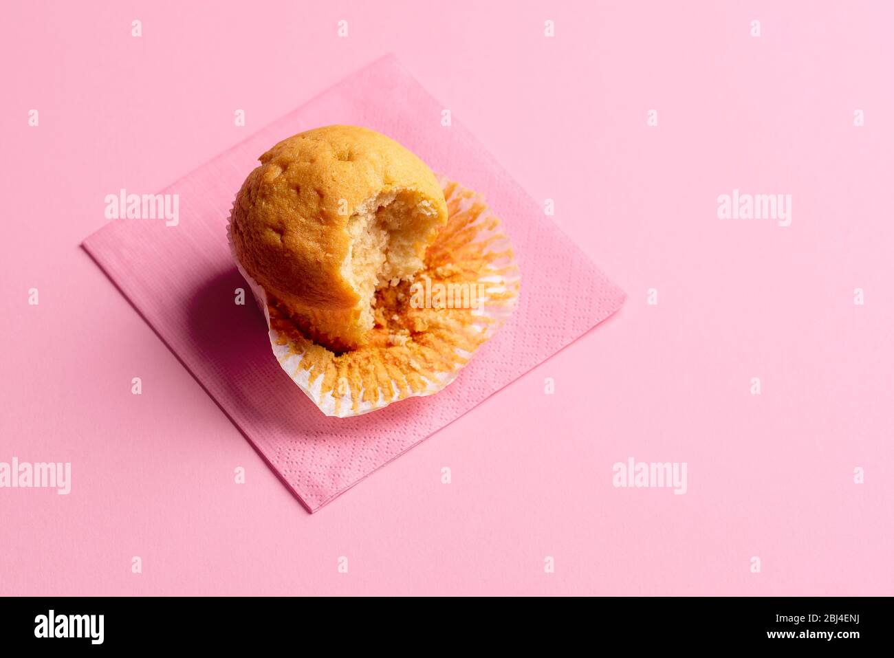 Muffin di base con un morso mancante su un tovagliolo, su uno sfondo rosa senza cuciture. Solo uno ha mangiato muffin. Cupcake parzialmente mangiato. Dessert popolare. Foto Stock