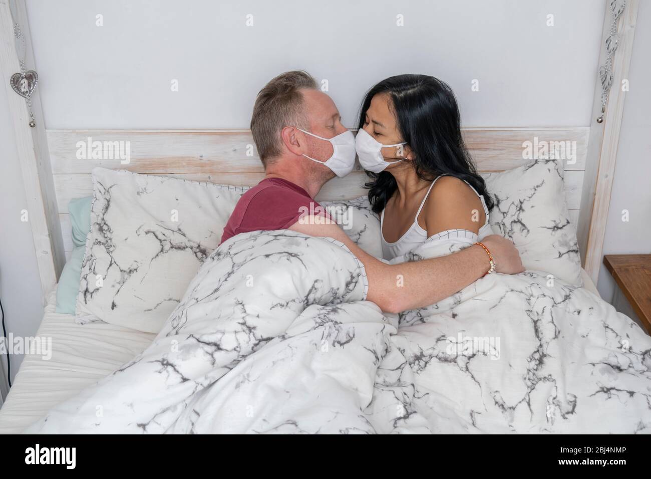 Coppia a letto indossando maschere viso baciando durante l'isolamento auto mentre in chiusura Foto Stock