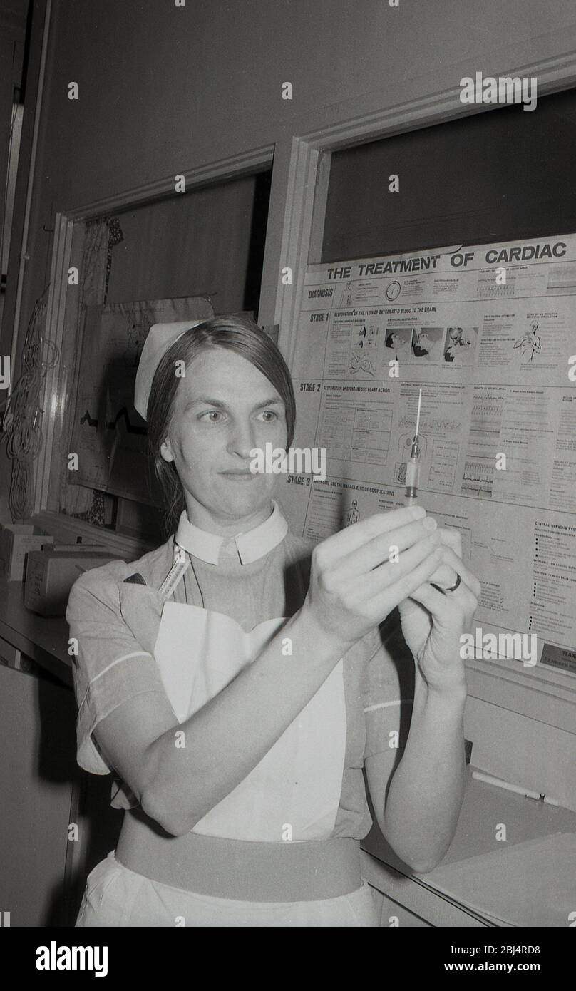 Anni '70, storica, un'infermiera femminile che tiene in su una siringa e ago medico, controllando il livello di liquido, Lewisham, Londra del sud, Inghilterra, Regno Unito. Le siringhe sono una pompa alternativa costituita da uno stantuffo (o pistone) che si adatta saldamente con un tubo sono utilizzate in medicina clinica per iniettare liquidi. Un grafico che descrive il trattamento dell'arresto cardico è su un tabellone dietro di lei. Foto Stock