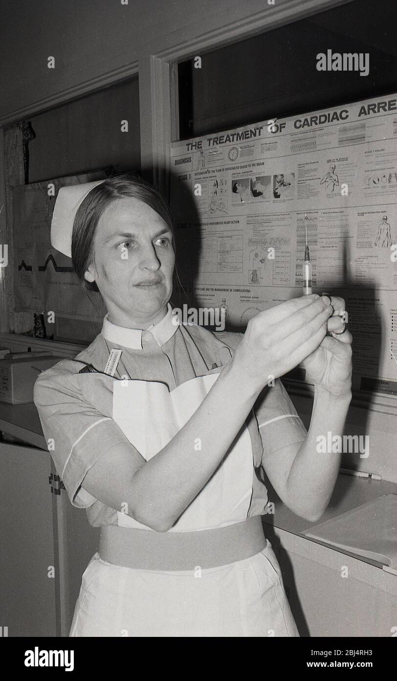 Anni '70, storica, un'infermiera femminile che tiene in mano una siringa e un ago medico di plastica, controllando il livello di liquido, Lewisham, Londra del Sud, Inghilterra, Regno Unito. Le siringhe sono una pompa alternativa costituita da uno stantuffo (o pistone) che si adatta saldamente con un tubo sono utilizzate in medicina clinica per iniettare liquidi. Un grafico che descrive il trattamento dell'arresto cardico è su un tabellone dietro di lei. Foto Stock