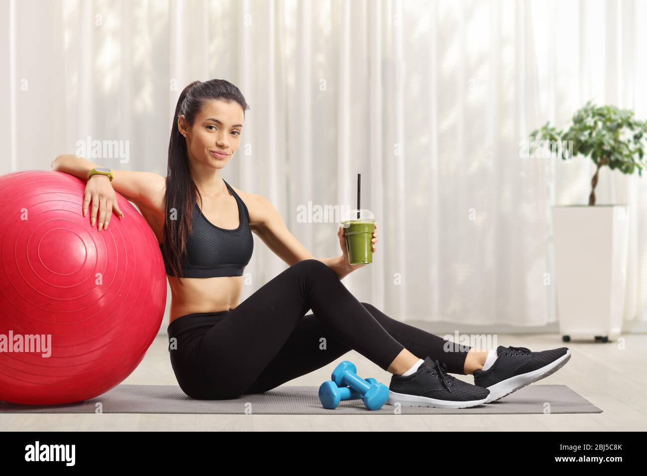 Adatta la donna seduta sul pavimento con una palla fitness e un sano frullato verde a casa Foto Stock