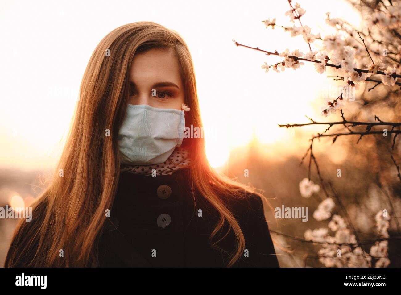 Ritratto di triste giovane donna ansiosa che indossa faccia maschera medica in piedi da albero fiorito nel parco al tramonto durante la primavera Foto Stock