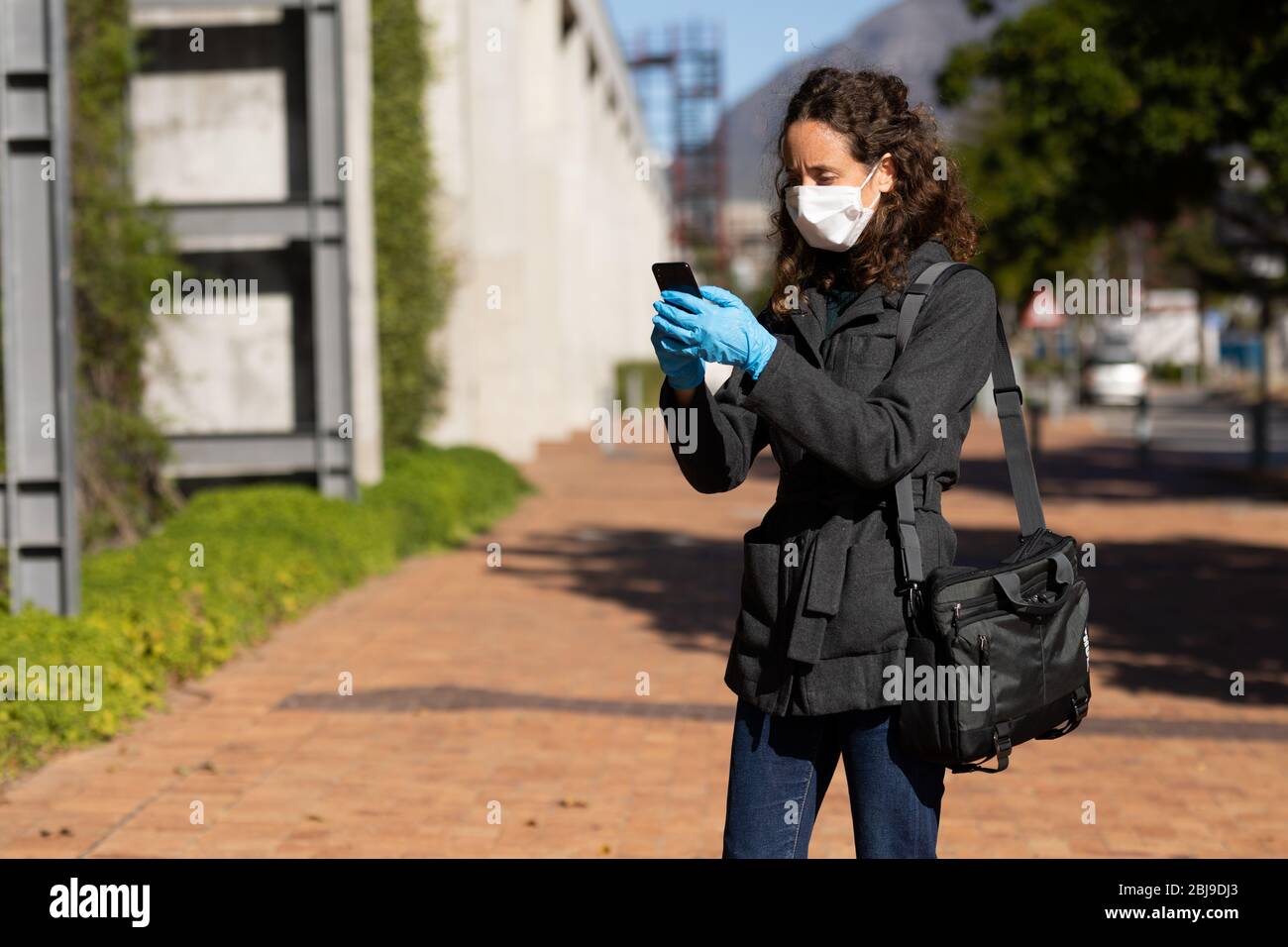 Donna caucasica che indossa una maschera protettiva contro il coronavirus fuori nelle strade, utilizzando il suo telefono Foto Stock