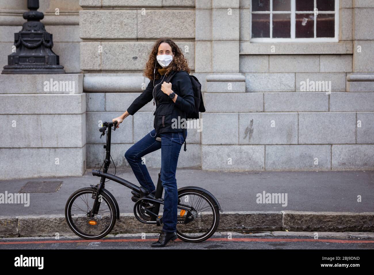 Donna caucasica che indossa una maschera protettiva e auricolari, e in bicicletta per le strade Foto Stock