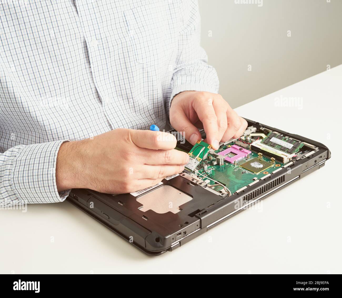 Uomo ripara il computer. Un tecnico dell'assistenza in camicia ripara il laptop, presso la scrivania bianca contro la parete bianca. Foto Stock