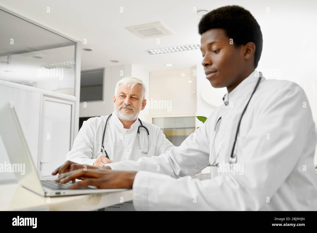 Terapisti multirazziali con stetoscopi sui colli che si pongono alla reception in ospedale. Medico africano maschio usando il laptop, fuoco selettivo di elde caucasico Foto Stock
