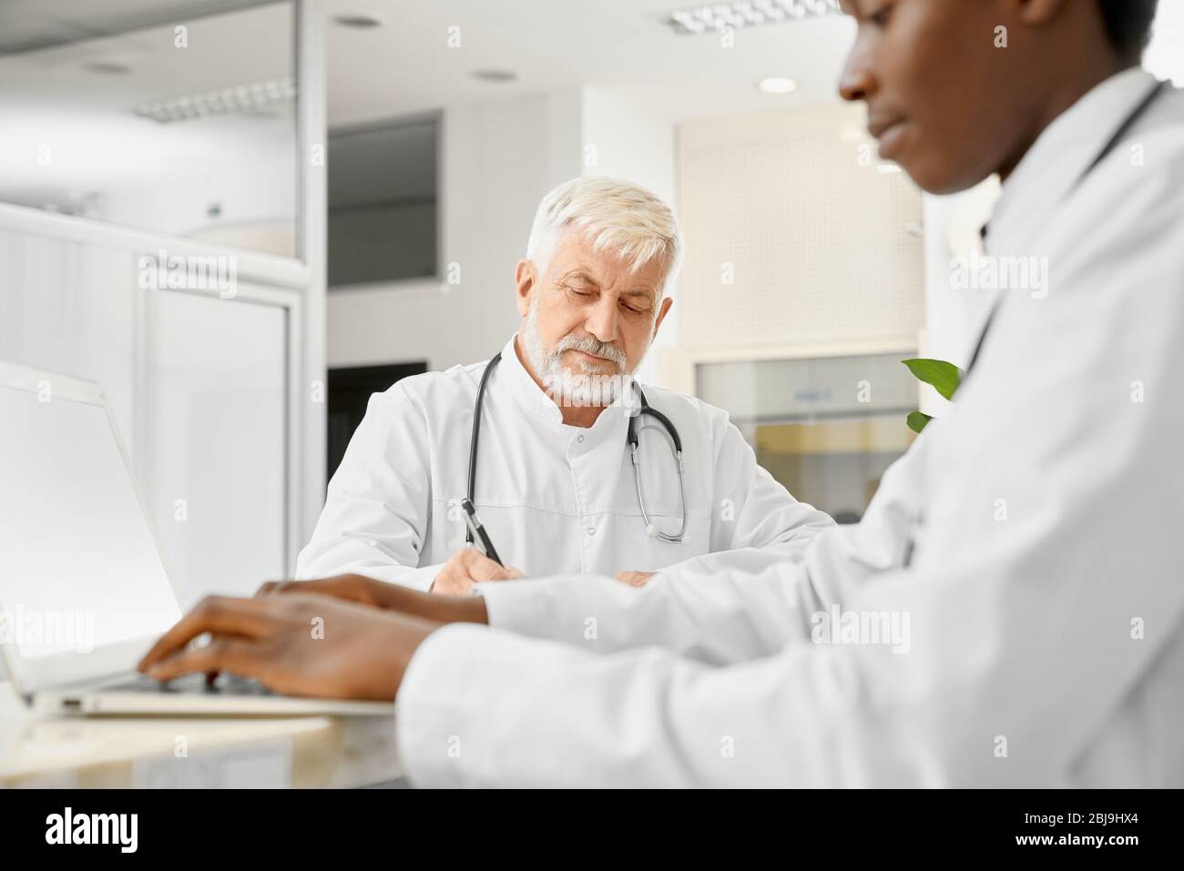 Terapisti con stetoscopi su colli che si pongono alla reception in ospedale. Medico africano maschio usando il laptop, fuoco selettivo del medico caucasico anziano ho Foto Stock