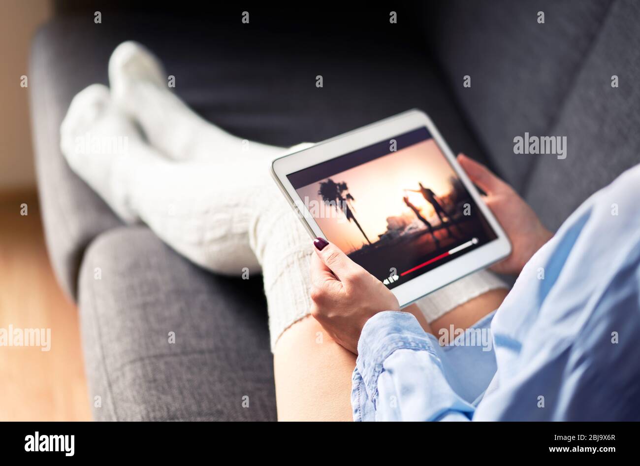 Donna che guarda serie tv o film in streaming con tablet sul divano che indossa calzini lunghi e accoglienti. Girl in streaming con dispositivo mobile e video on demand. Foto Stock