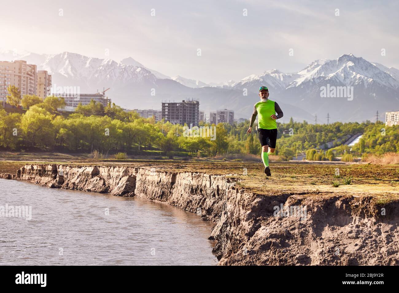 Uomo anziano in esecuzione nei pressi del fiume con edifici e lo sfondo di montagna al mattino. Uno stile di vita sano concetto Foto Stock