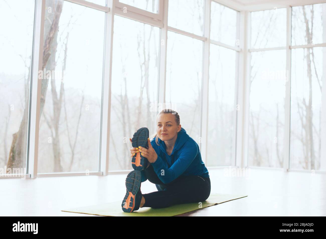 La donna concentrata sta facendo un po' di stretching sul pavimento in una stanza piena di luce e finestre. Foto Stock