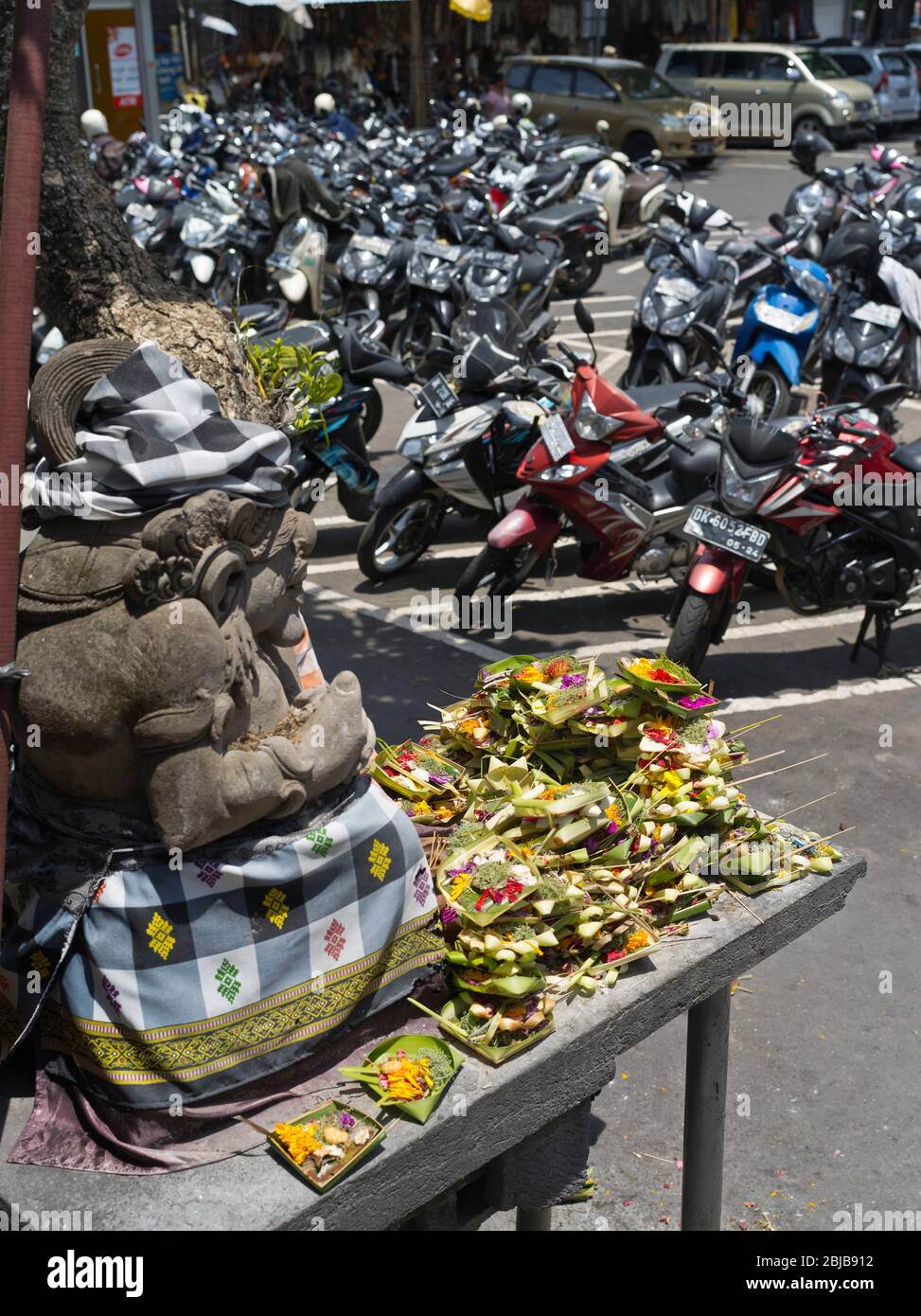 dh Balinese mercato carpark BALI INDONESIA ASIA statua indù custode offerte nel parcheggio scooter parcheggiato offerte di cibo Foto Stock