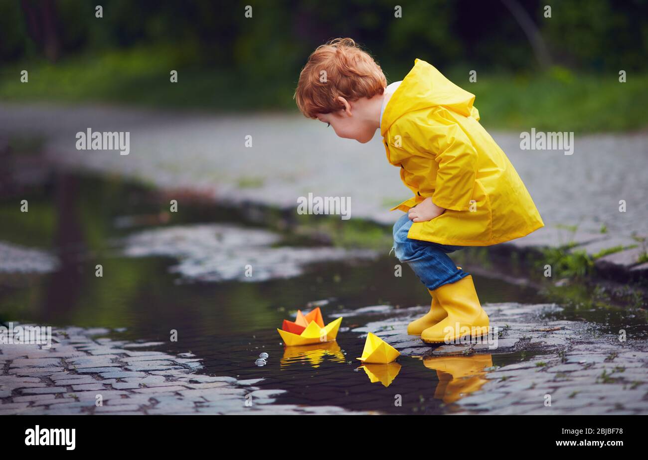 carino bambino ragazzo lanciare barche di carta in primavera, indossando impermeabile e stivali di gomma Foto Stock