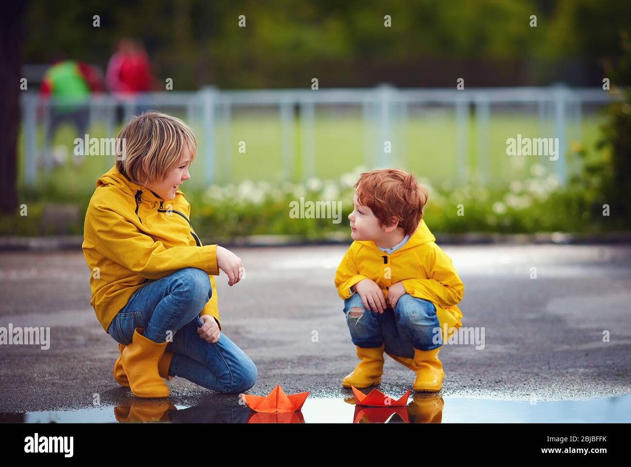 ragazzi carini, fratelli che si divertono insieme, lanciando le barche di carta in pozzanghere primaverili, indossando impermeabili e stivali di gomma Foto Stock