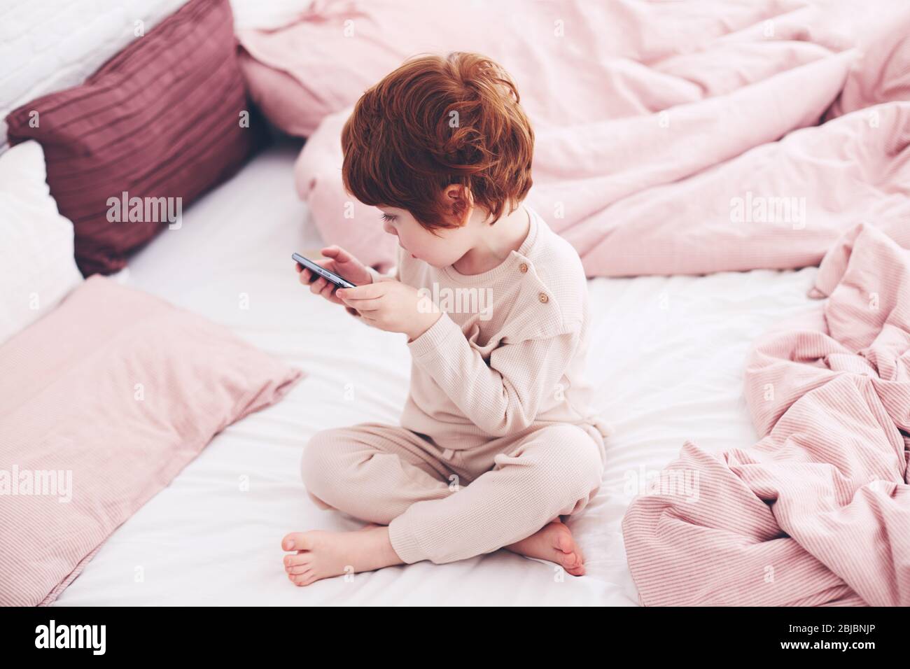 cute redhead bambino ragazzo in pajama giocare video giochi al telefono in camera da letto al mattino Foto Stock