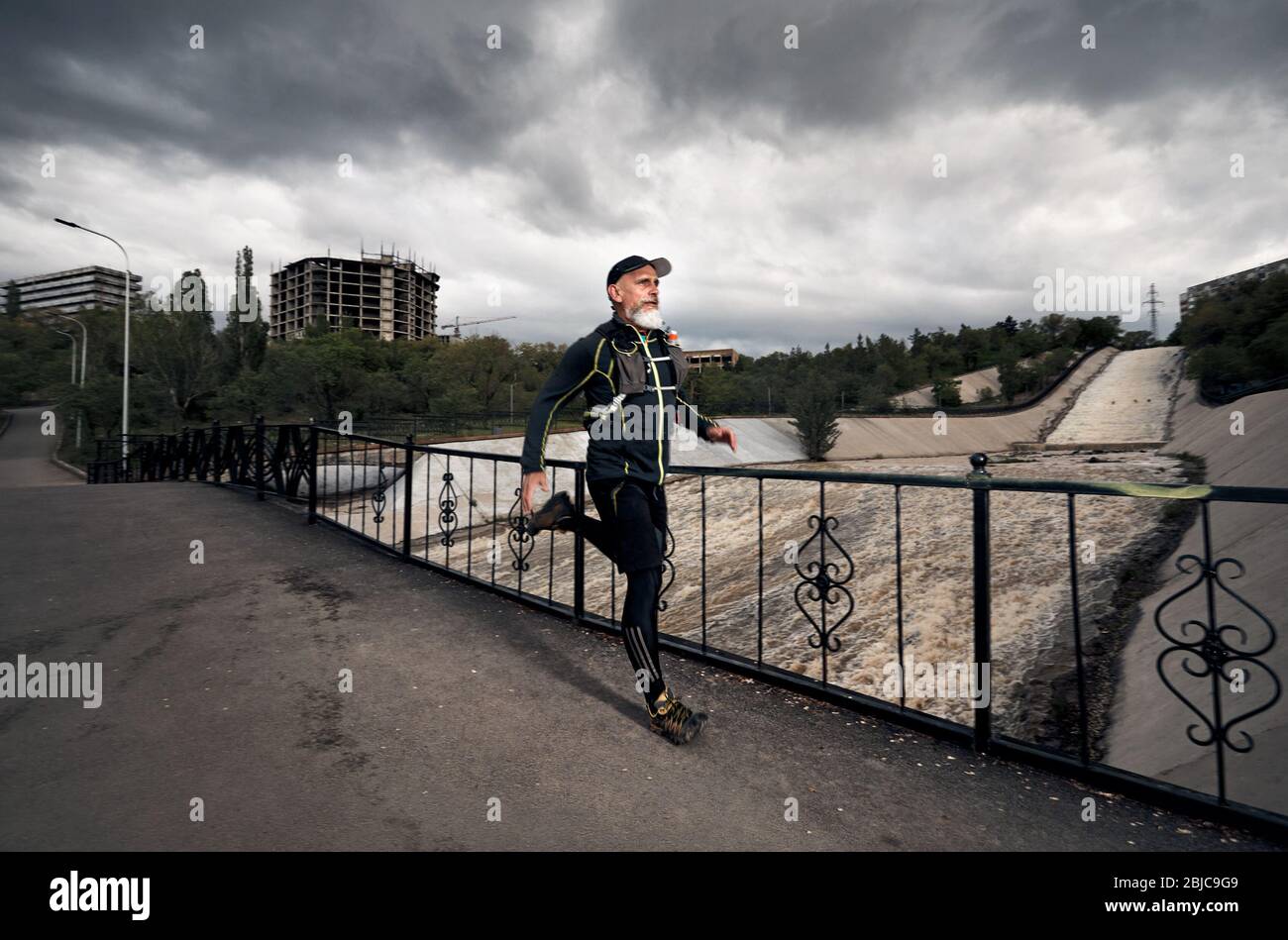 Uomo con barba grigia in costume nero e zainetto in esecuzione sul ponte che attraversa il fiume sporco a sfondo con cielo nuvoloso Foto Stock