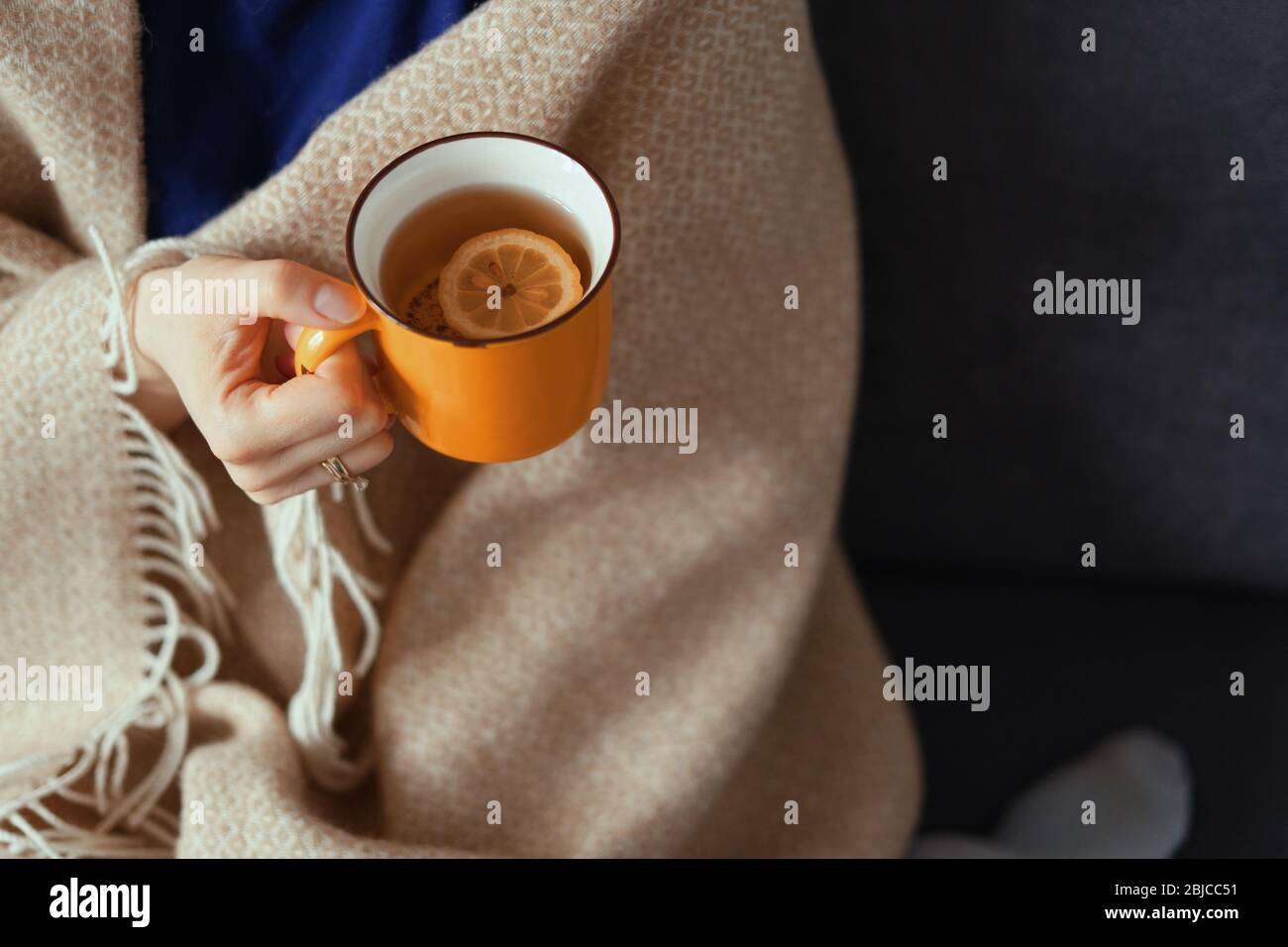 Mano della donna che tiene una tazza gialla di tè con limone in una giornata fredda. Momenti comodi, coperta di lana Foto Stock