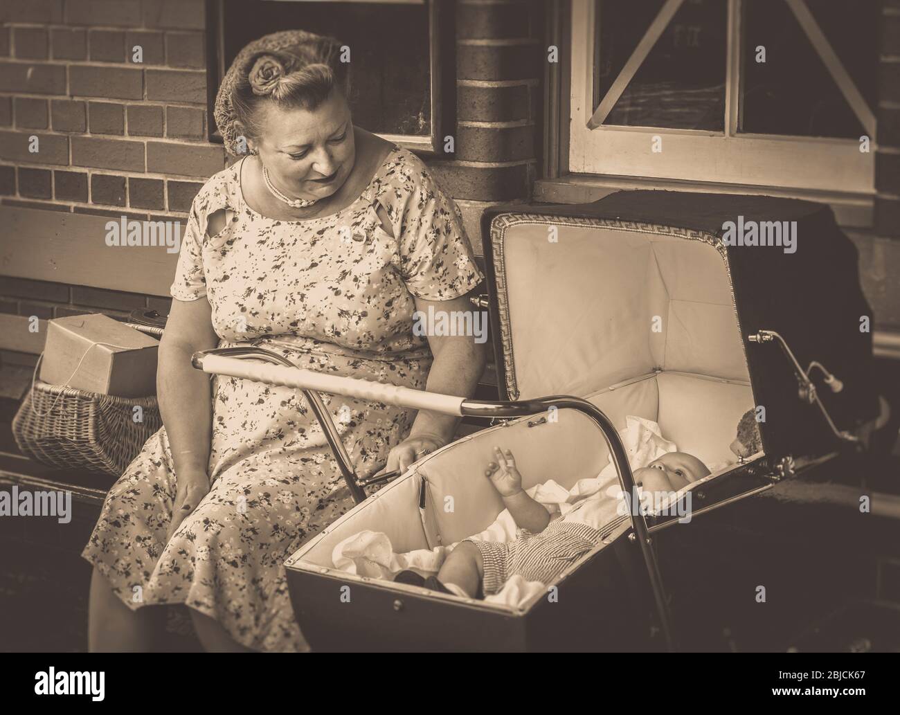 Seppia vista frontale della donna degli anni '40 in moda anni '40 con il bambino in vintage pram, stazione ferroviaria patrimonio, WWII evento estivo in tempo di guerra, Regno Unito. Foto Stock