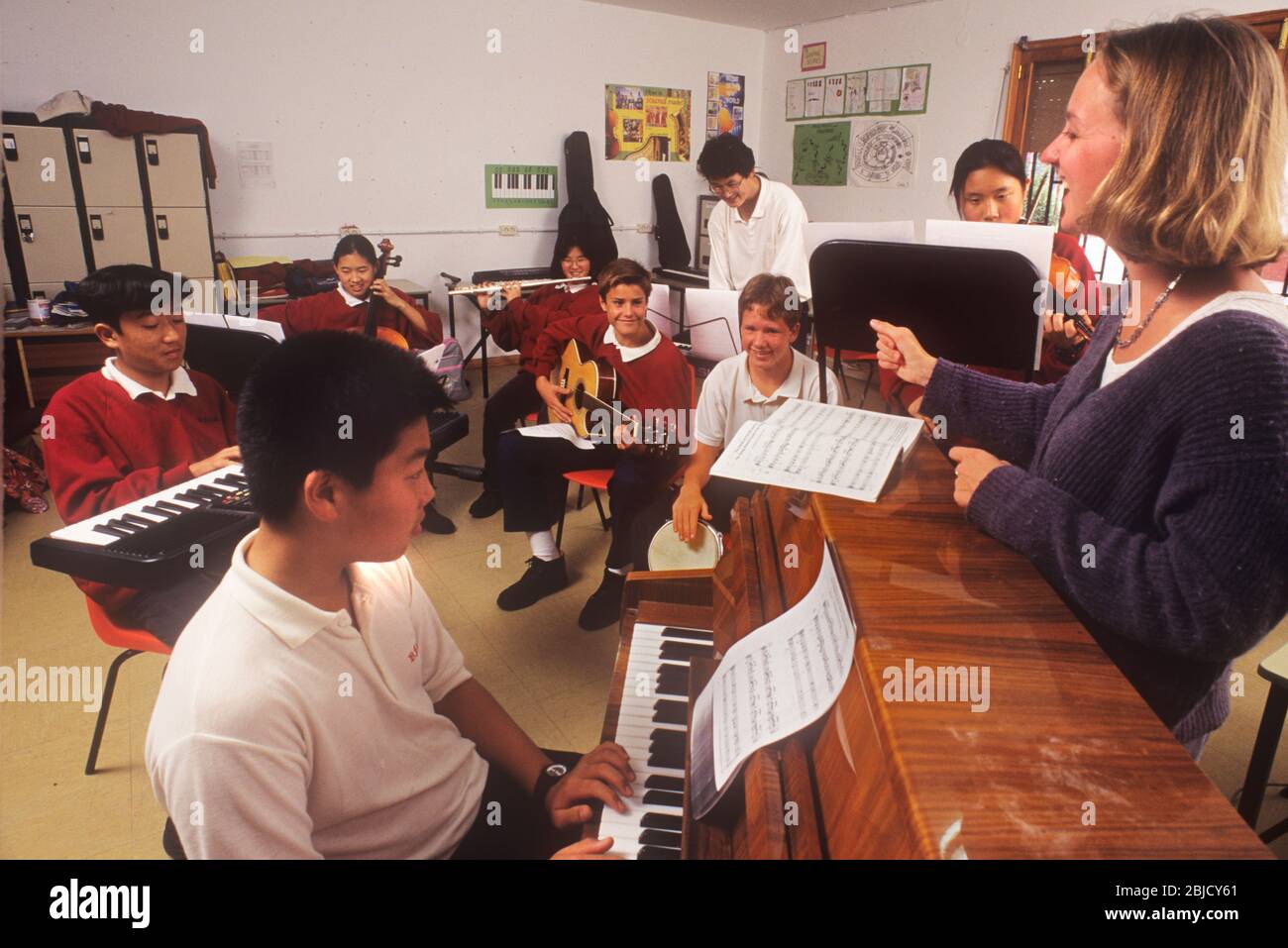 Classe musicale in aula scolastica con alunni 13-15 anni che suonano vari strumenti con un'entusiasta insegnante femminile che conduce una prova Foto Stock