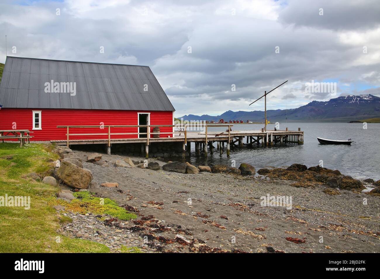 Vista sul fiordo con un molo, tradizionale edificio in legno rosso, barca da pesca, belle montagne e isole sullo sfondo, Eskifjordur, Islanda Foto Stock