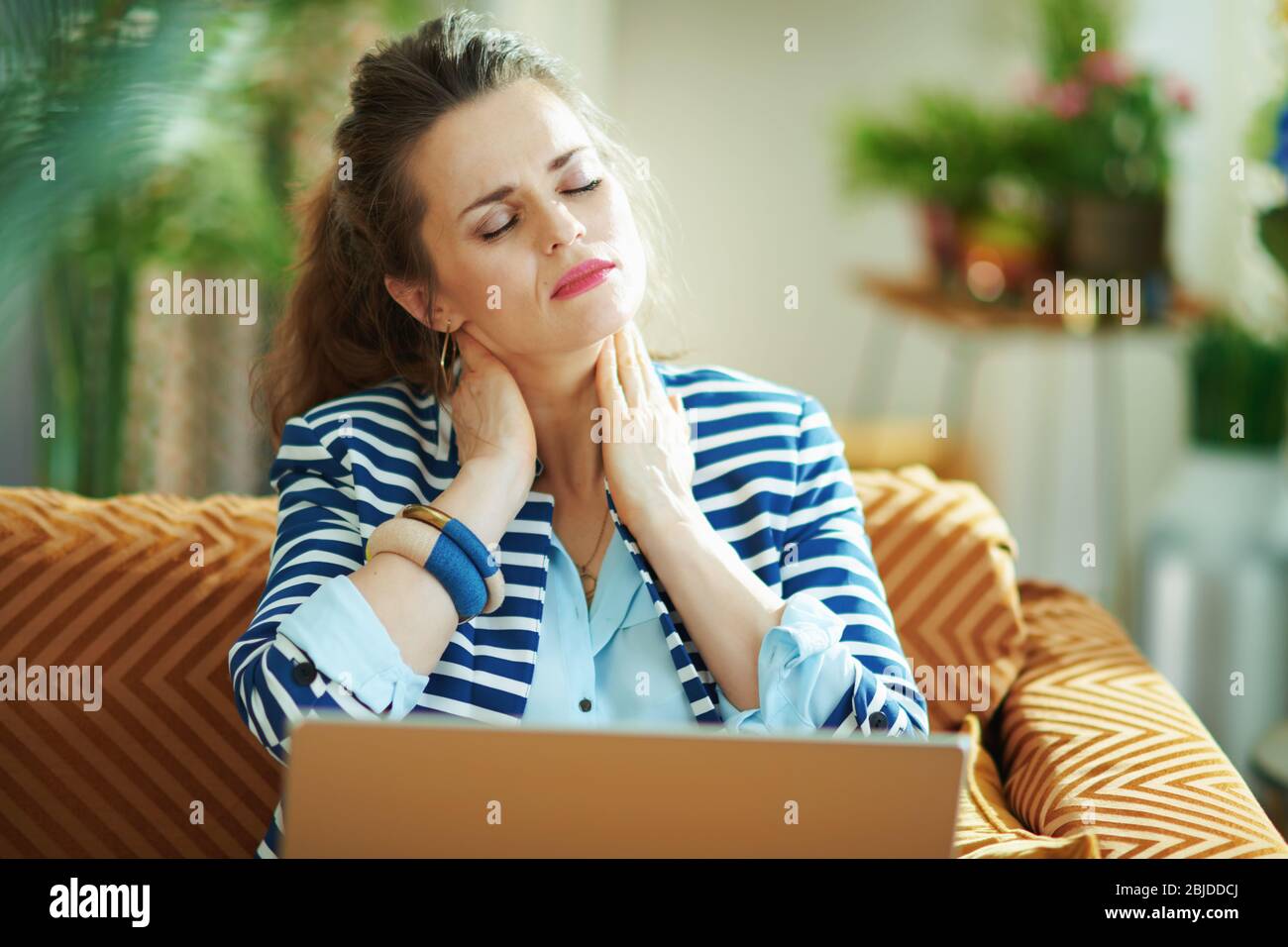 donna stressata in blusa blu e giacca a righe con dolore al collo seduto sul divano con il laptop a casa moderna in sole. Foto Stock