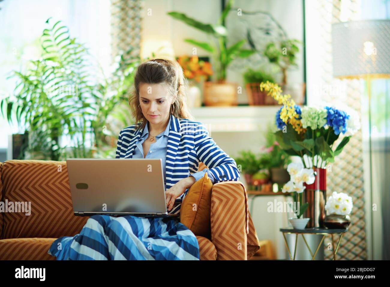 elegante donna in blusa blu e giacca a righe seduta sul divano con laptop nella casa moderna in giorno di sole. Foto Stock
