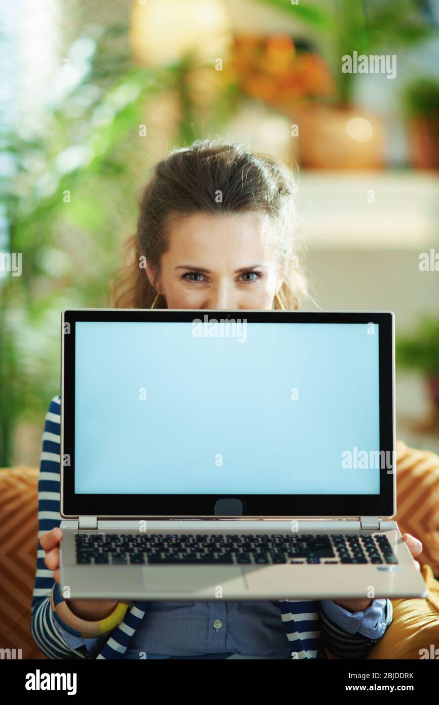 giovane donna in blusa blu e giacca a righe in casa moderna in una giornata di sole seduta sul divano e mostrando il laptop schermo vuoto. Foto Stock