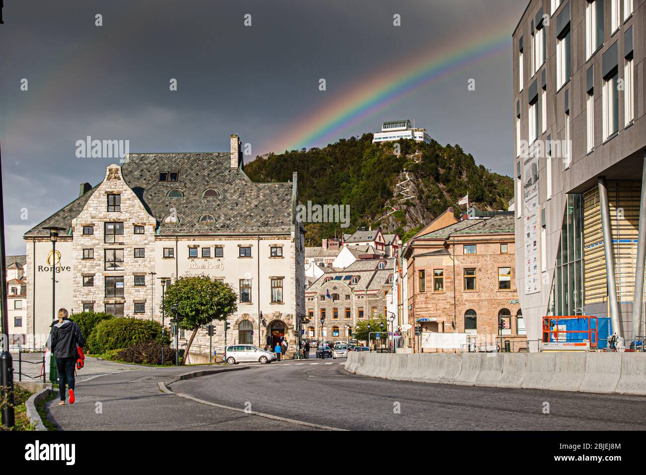 Un arcobaleno si inarca qui sopra la vecchia città di Ålesund, in Norvegia, verso la collina del punto di riferimento locale di Aksla. Sulla sua cima si trova forse la piattaforma panoramica Fjellstua, raggiungibile tramite le cosiddette 418 scale. In primo piano, un passante cammina lungo la strada bagnata dalla pioggia. Foto Stock