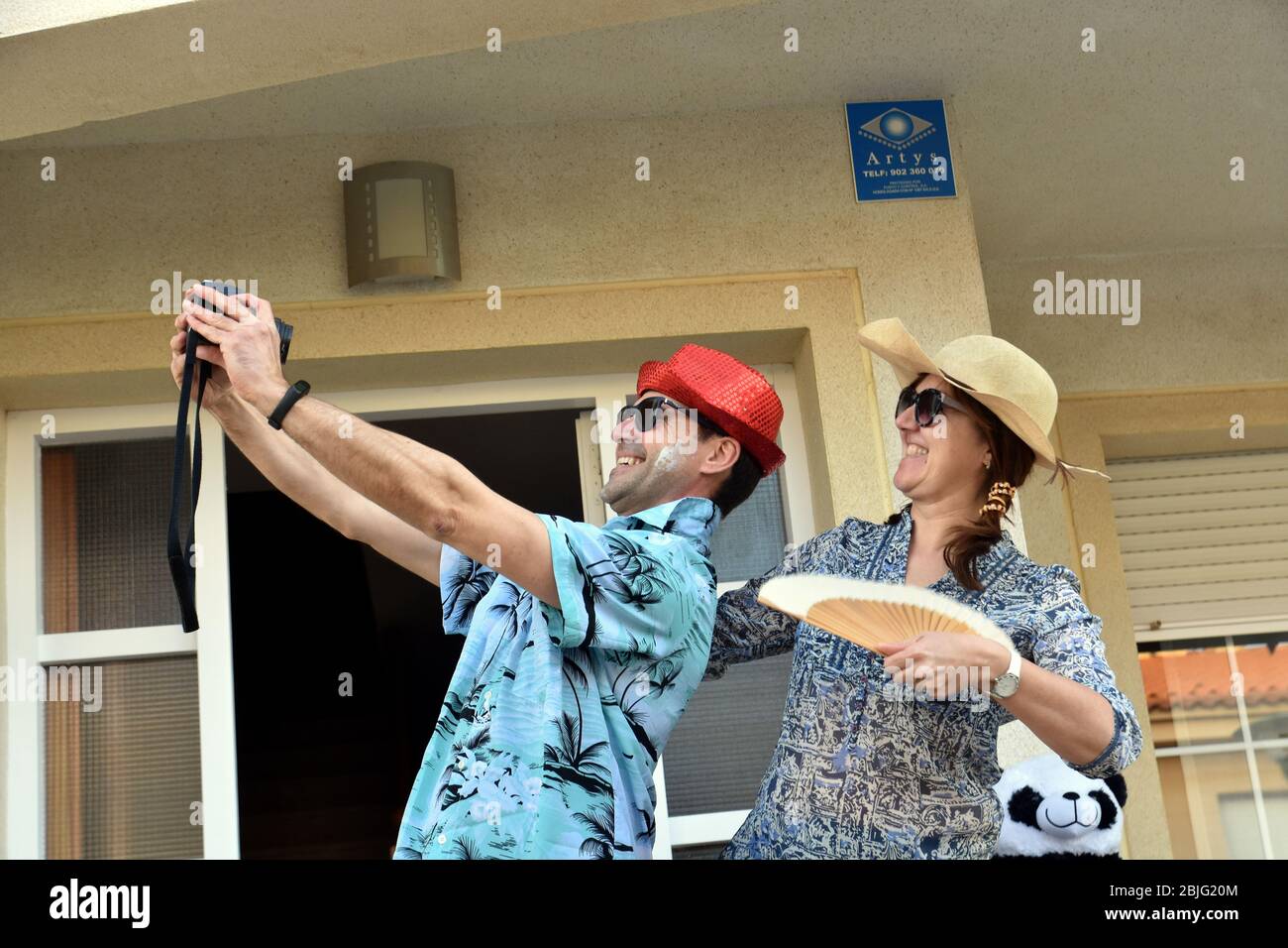 Una coppia all'ingresso della loro casa mascherata come turisti stranieri posa per un selfie per incoraggiarli durante il confinamento tra Crisis.Residents Coronavirus di Mas d'en quartiere di Gual eseguire e applausi alle 8 pm per incoraggiare i loro vicini durante il confinamento tra la crisi del coronavirus. Ogni giorno scelgono un tema, Musica degli anni '80, Fiera di aprile, oggi hanno fatto uno spettacolo travestito da turisti con le loro valigie e infradito con calze. Foto Stock