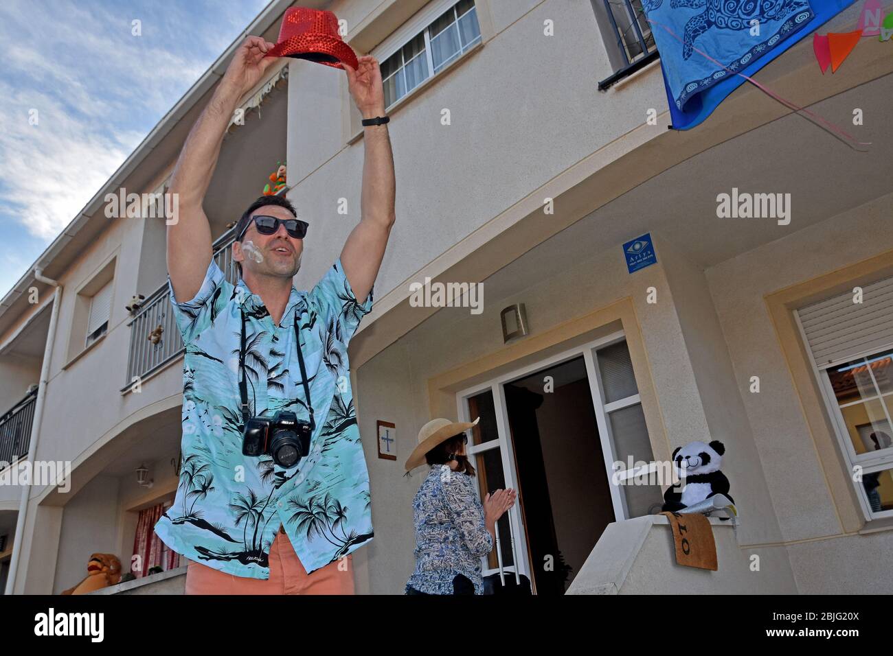 Un uomo vestito come un turista con una crema solare applicata sul suo viso saluta i suoi vicini sulla terrazza della sua casa per incoraggiarli durante il confinamento tra Crisis.Residents Coronavirus di Mas d'en quartiere di Gual eseguire e applausi alle 8 pm per incoraggiare i loro vicini durante il confinamento in mezzo alla crisi del coronavirus. Ogni giorno scelgono un tema, Musica degli anni '80, Fiera di aprile, oggi hanno fatto uno spettacolo travestito da turisti con le loro valigie e infradito con calze. Foto Stock