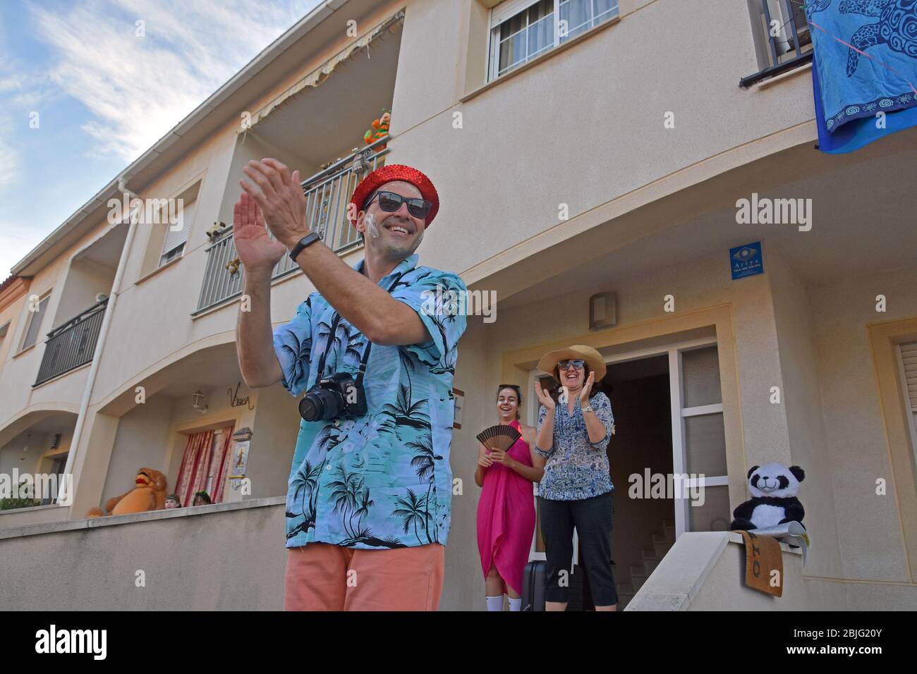 Un uomo vestito come un turista con una crema solare applicata sulla sua faccia applaudes i suoi vicini sulla terrazza della sua casa per incoraggiarli durante il confinamento tra Crisis.Residents Coronavirus di Mas d'en quartiere di Gual eseguire e applausi alle 8 pm per incoraggiare i loro vicini durante il confinamento in mezzo alla crisi del coronavirus. Ogni giorno scelgono un tema, Musica degli anni '80, Fiera di aprile, oggi hanno fatto uno spettacolo travestito da turisti con le loro valigie e infradito con calze. Foto Stock