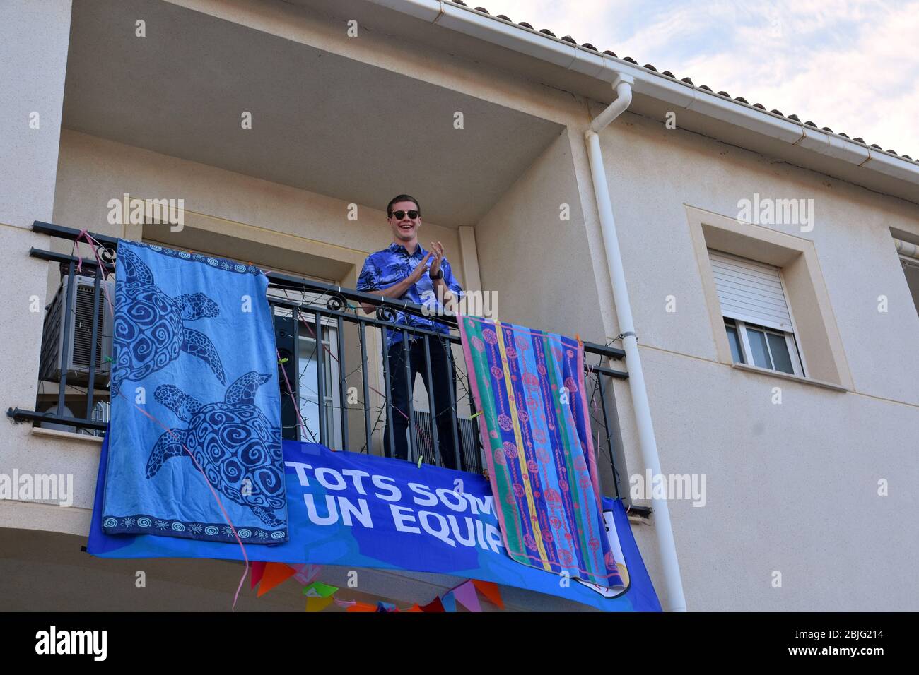 Un uomo applaude i suoi vicini sulla terrazza della sua casa per incoraggiarli durante il confinamento tra Crisis.Residents Coronavirus Crisis.Residents di Mas d'en Gual quartiere eseguire e applausi alle 8 pm per incoraggiare i loro vicini durante il confinamento in mezzo alla crisi del coronavirus. Ogni giorno scelgono un tema, Musica degli anni '80, Fiera di aprile, oggi hanno fatto uno spettacolo travestito da turisti con le loro valigie e infradito con calze. Foto Stock