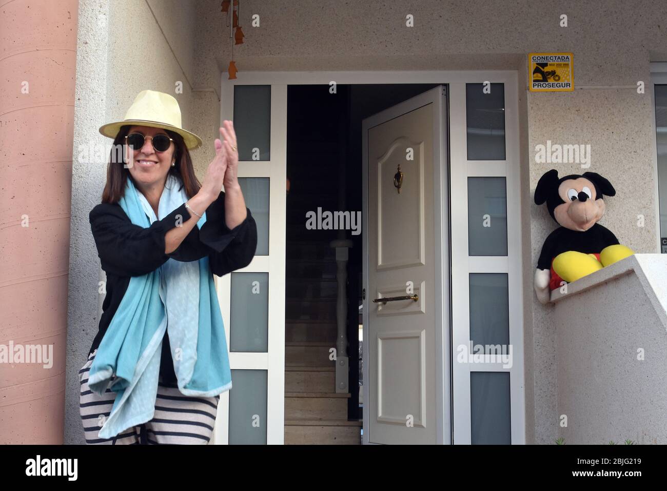 Una donna in un cappello applaudes i suoi vicini sulla terrazza della sua casa per incoraggiarli durante il confinamento tra Crisis.Residents Coronavirus Crisis.Residents di Mas d'en Gual quartiere eseguire e applausi alle 8 pm per incoraggiare i loro vicini durante il confinamento tra la crisi del coronavirus. Ogni giorno scelgono un tema, Musica degli anni '80, Fiera di aprile, oggi hanno fatto uno spettacolo travestito da turisti con le loro valigie e infradito con calze. Foto Stock