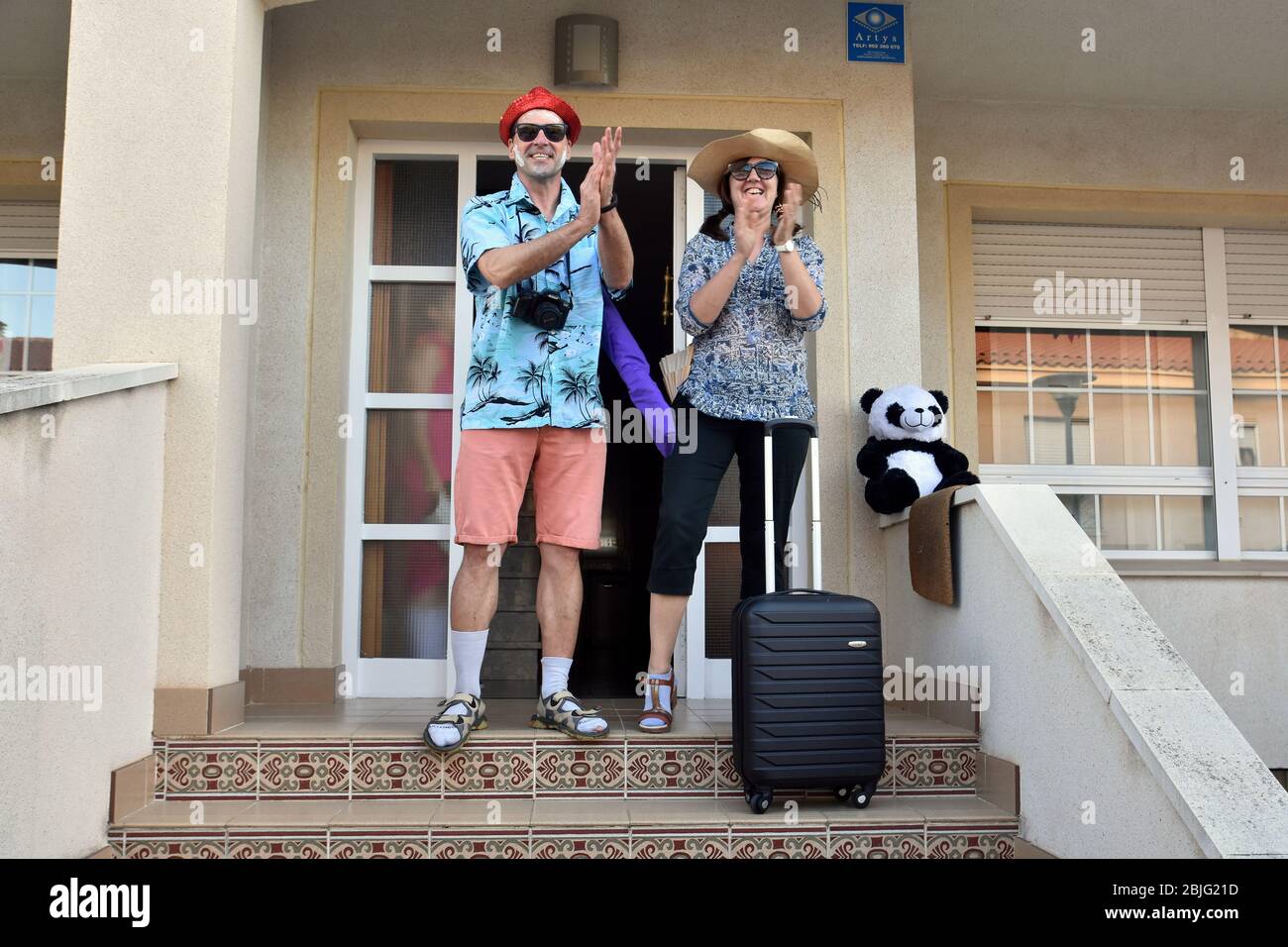Una coppia travestita come turisti stranieri con valigie applaudono i loro vicini sulla terrazza della loro casa per incoraggiarli durante il confinamento tra Crisis.Residents Coronavirus di Mas d'en quartiere di Gual eseguire e applausi alle 8 pm per incoraggiare i loro vicini durante il confinamento tra la crisi del coronavirus. Ogni giorno scelgono un tema, Musica degli anni '80, Fiera di aprile, oggi hanno fatto uno spettacolo travestito da turisti con le loro valigie e infradito con calze. Foto Stock