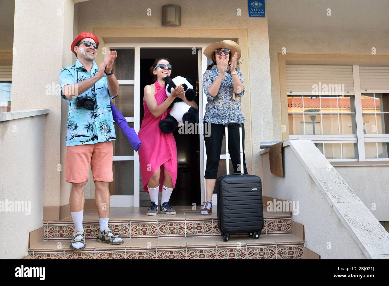 Una famiglia travestita come turisti stranieri con valigie applaudono i loro vicini sulla terrazza della loro casa per incoraggiarli durante il confinamento tra Cionavirus Crisis.Residents di Mas d'en Gual quartiere eseguire e applausi alle 8 pm per incoraggiare i loro vicini durante il confinamento tra la crisi del coronavirus. Ogni giorno scelgono un tema, Musica degli anni '80, Fiera di aprile, oggi hanno fatto uno spettacolo travestito da turisti con le loro valigie e infradito con calze. Foto Stock