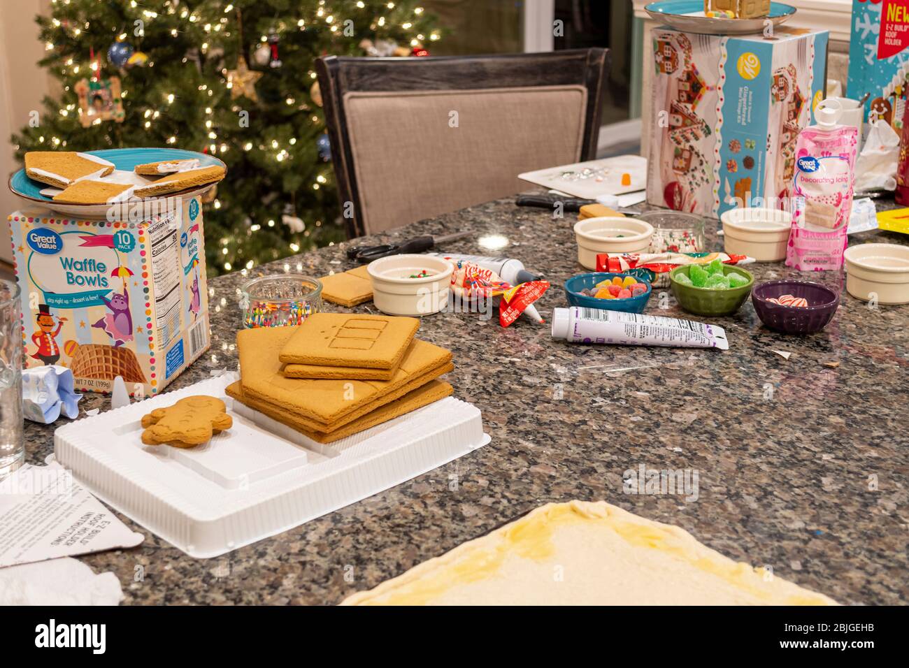 Vari ingredienti della casa di Gingerbread sono disposti su un bancone della cucina di granito con un albero di Natale illuminato dietro. Foto Stock