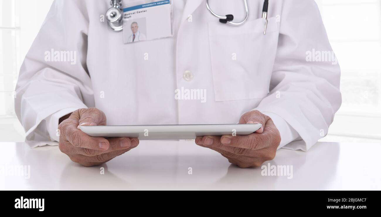 Primo piano di un medico che tiene un computer tablet nelle sue mani mentre si siede alla sua scrivania in un ambiente di ufficio High Key. L'attenzione è rivolta a tablet e mani, Foto Stock